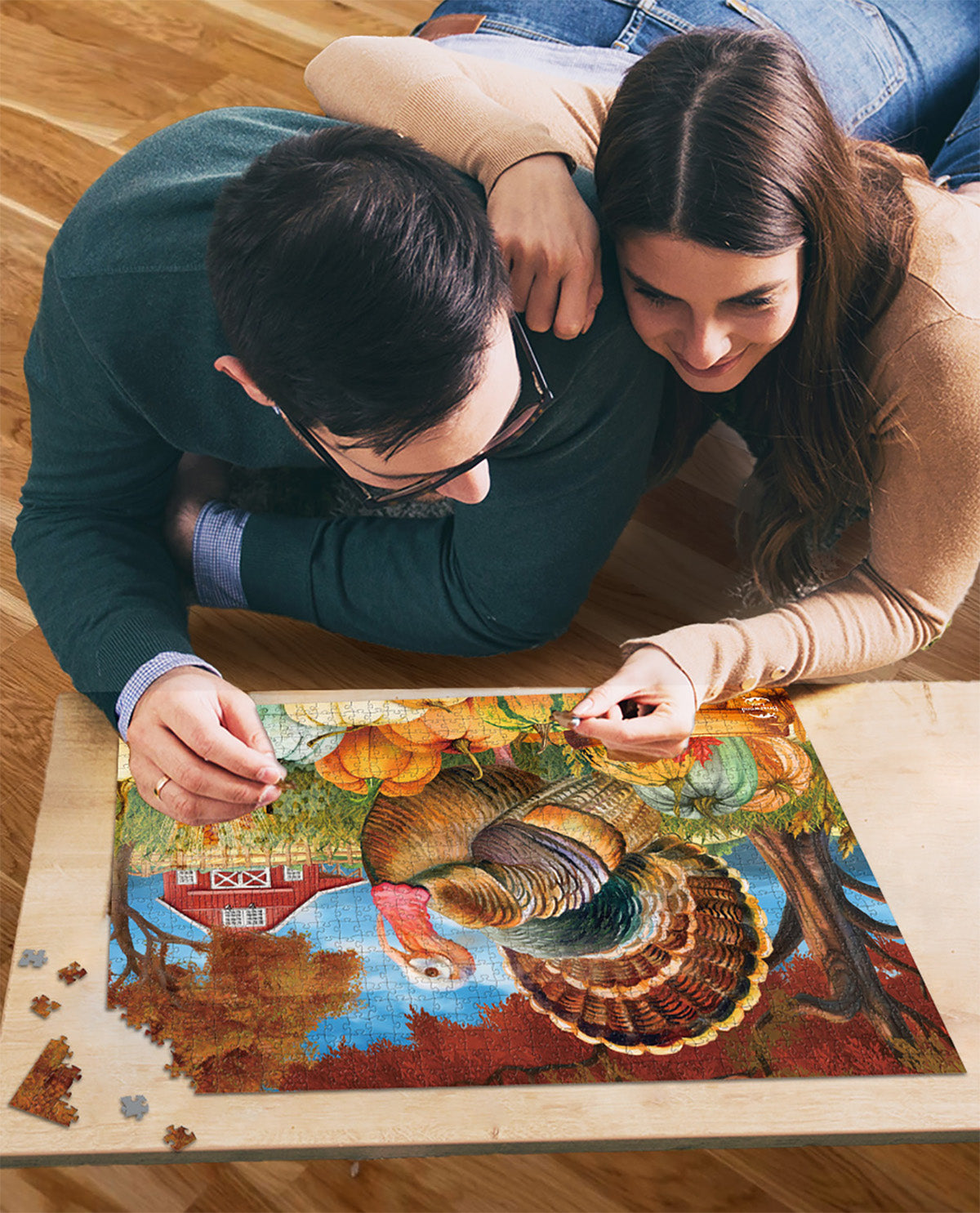 A couple lying on a wooden floor looking at a completed jigsaw puzzle of a Thanksgiving turkey in a autumn farm scene.