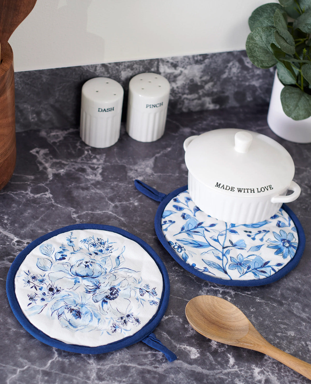A set of blue and white floral kitchen accessories on a dark marble countertop, including two round potholders, a matching pot holder with a white lidded casserole dish, a wooden spoon, and salt and pepper shakers in the background.