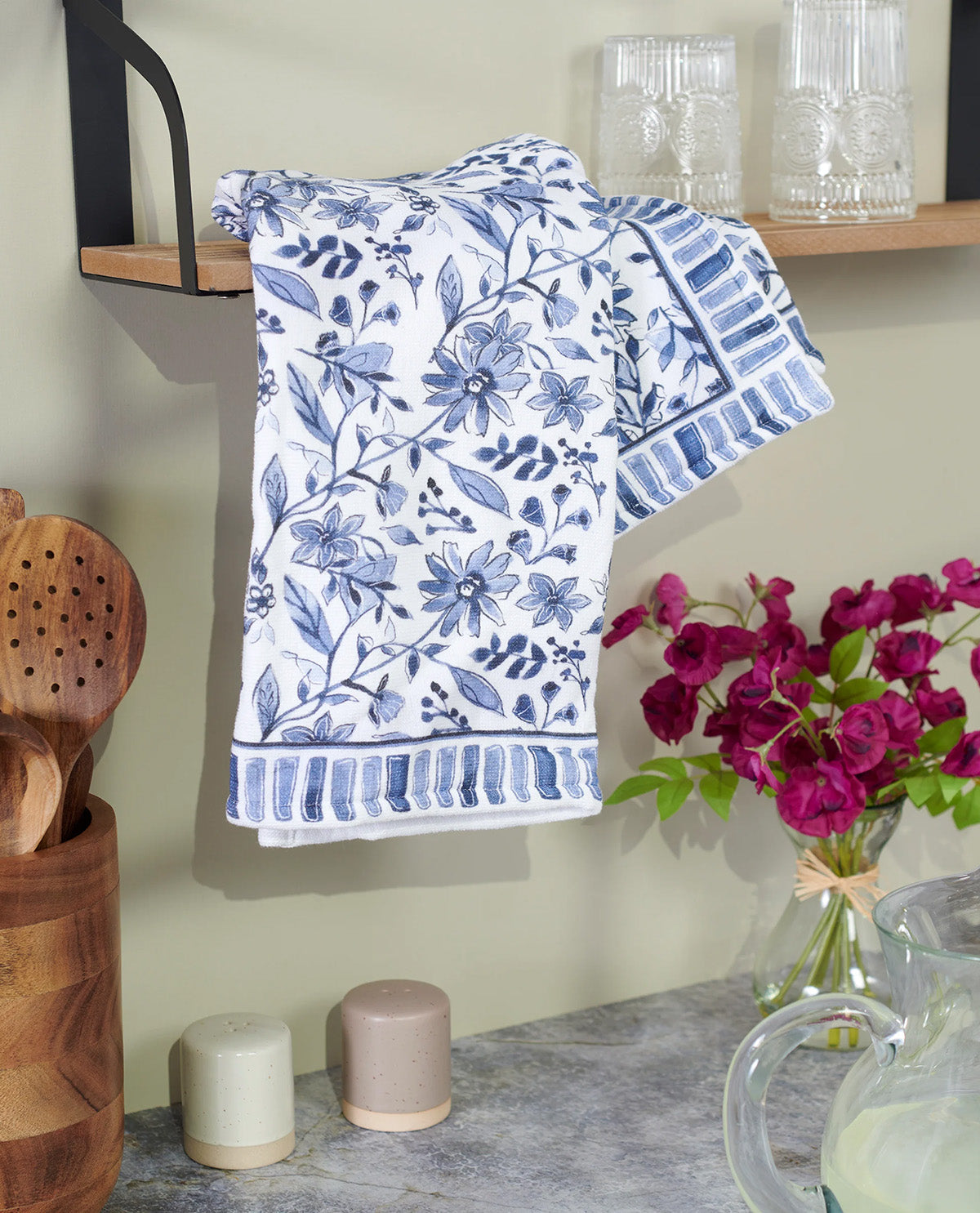 A white kitchen towel with a blue floral and striped pattern hanging on a wall hook in a kitchen setting, with a shelf, glassware, flowers, and countertop items visible.