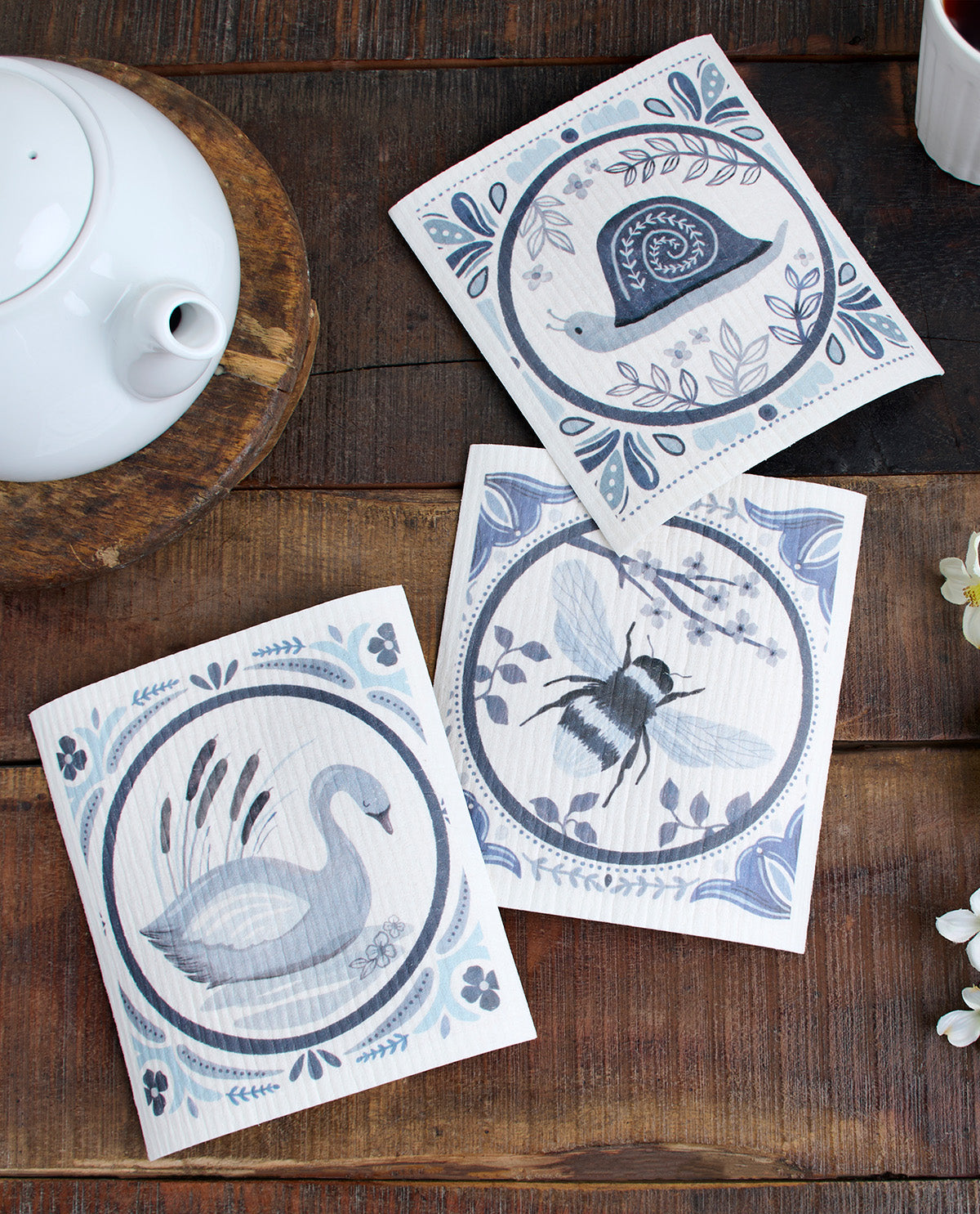 Three square decorative paper napkins with blue and gray nature illustrations (a swan, a bee, and a snail) arranged on a dark wooden table next to a white teapot.