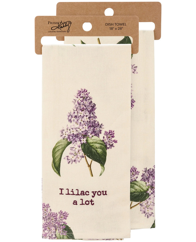 A cream-colored dish towel with a purple lilac floral print and the text 'I'll lac you a lot' in purple, hanging from a brown kraft paper hanger. A second identical towel is partially visible behind it.