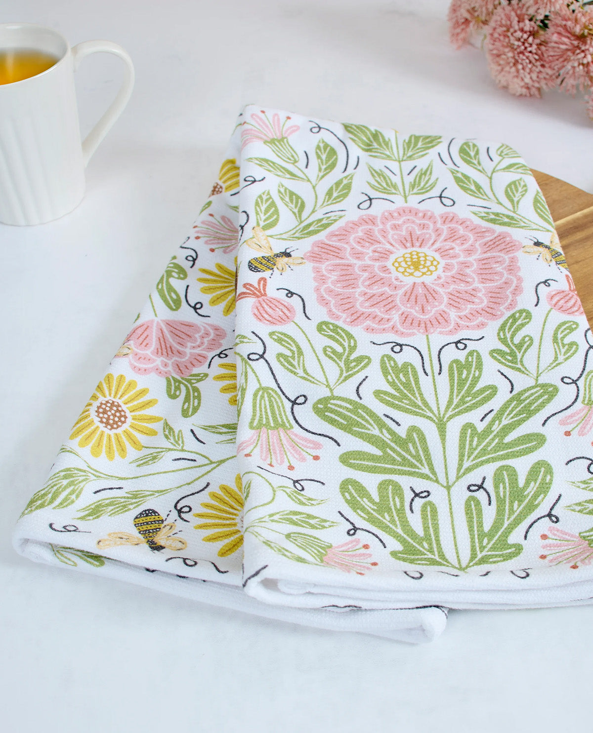 A folded white kitchen towel with a colorful floral and bee pattern, styled on a light surface next to a white mug and pink flowers.