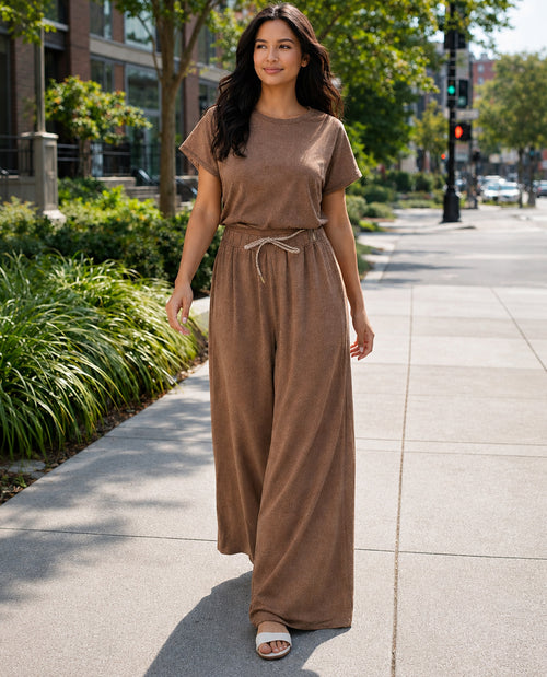 A woman wearing a matching two-piece lounge set in a warm taupe-brown color stands on a sunny city sidewalk. The set includes a short-sleeve crewneck top and matching wide-leg pants with a drawstring waist.