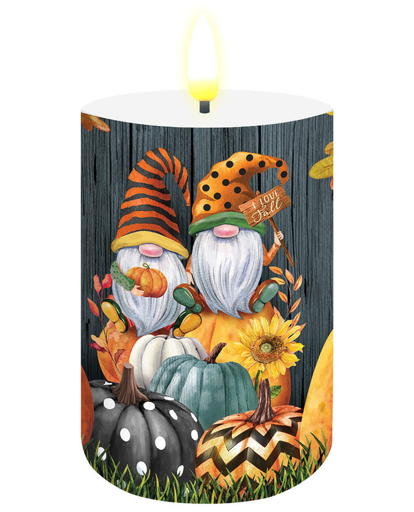 A lit white pillar candle with a printed autumn gnomes design, featuring two gnomes with orange and yellow hats, pumpkins, sunflowers, and a dark wood plank background.