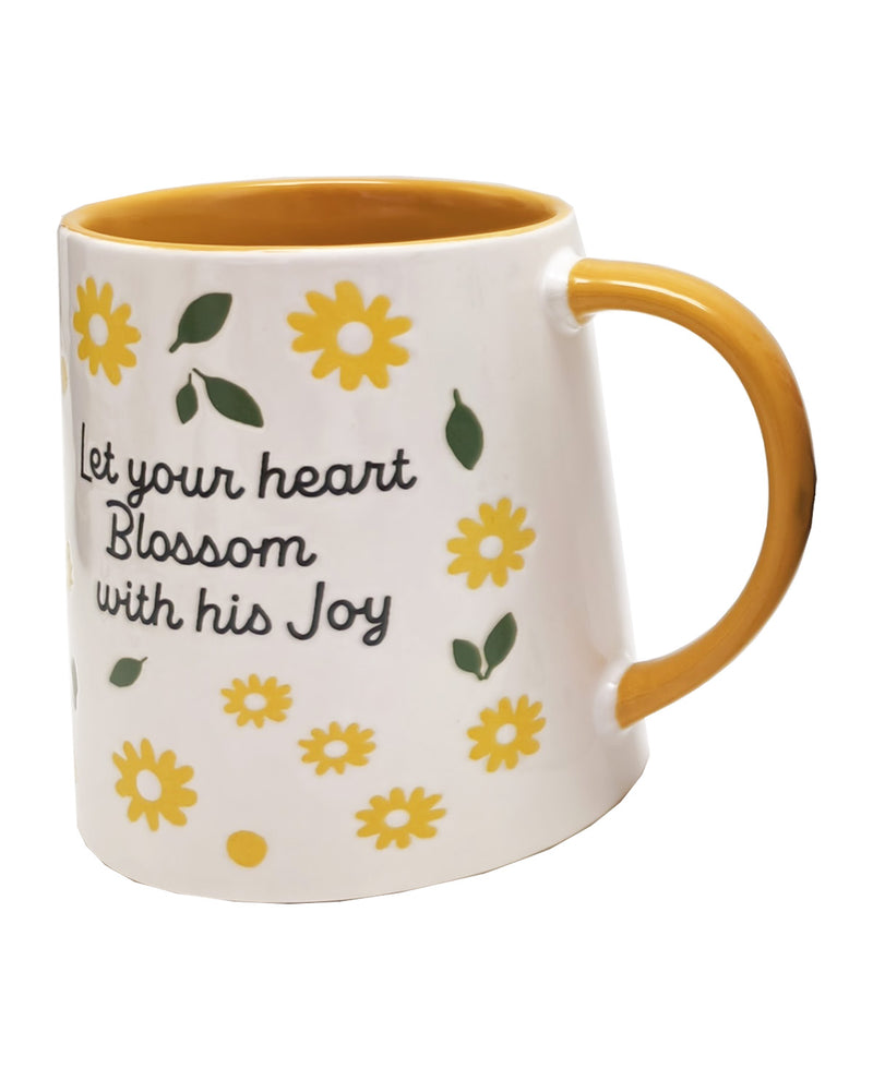 A white ceramic mug with a mustard yellow handle and interior, decorated with yellow flowers, green leaves, and the printed phrase 'Let your heart Blossom with his Joy' in black script.