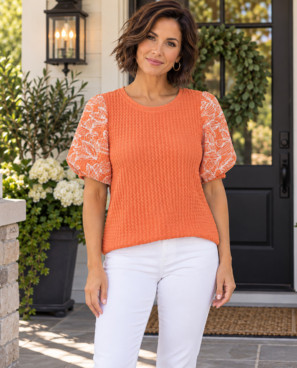 A woman stands on a front porch wearing a textured orange short-sleeve top with white lace flutter sleeves and white pants.