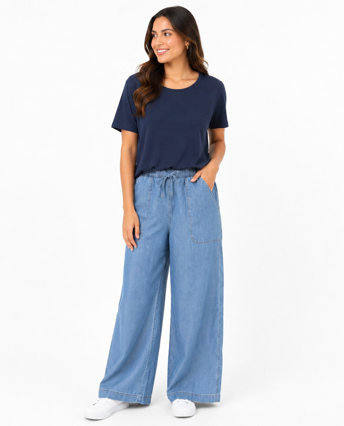 A woman modeling a navy blue short-sleeve crewneck t-shirt and light blue wide-leg denim pants with white sneakers, standing against a plain white studio background.