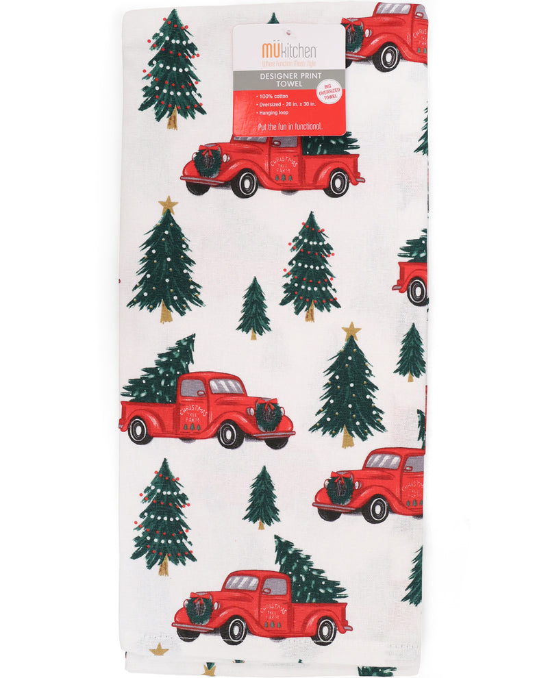 A folded white fabric item, likely a tea towel or bandana, featuring a repeating pattern of vintage red pickup trucks carrying Christmas trees and green evergreen trees. A paper label for 'musiken Designer Print' is attached at the top.