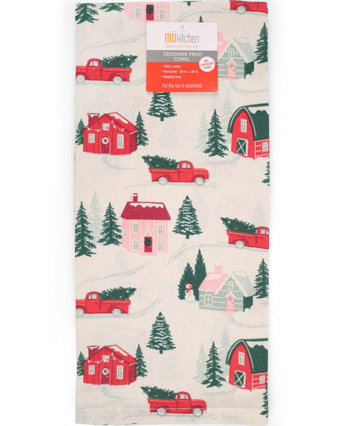 A folded kitchen towel with a festive Christmas print featuring red pickup trucks carrying Christmas trees, red and pink houses, green pine trees, and red barns on an off-white background. A paper label from 'mukitchen' is attached at the top.