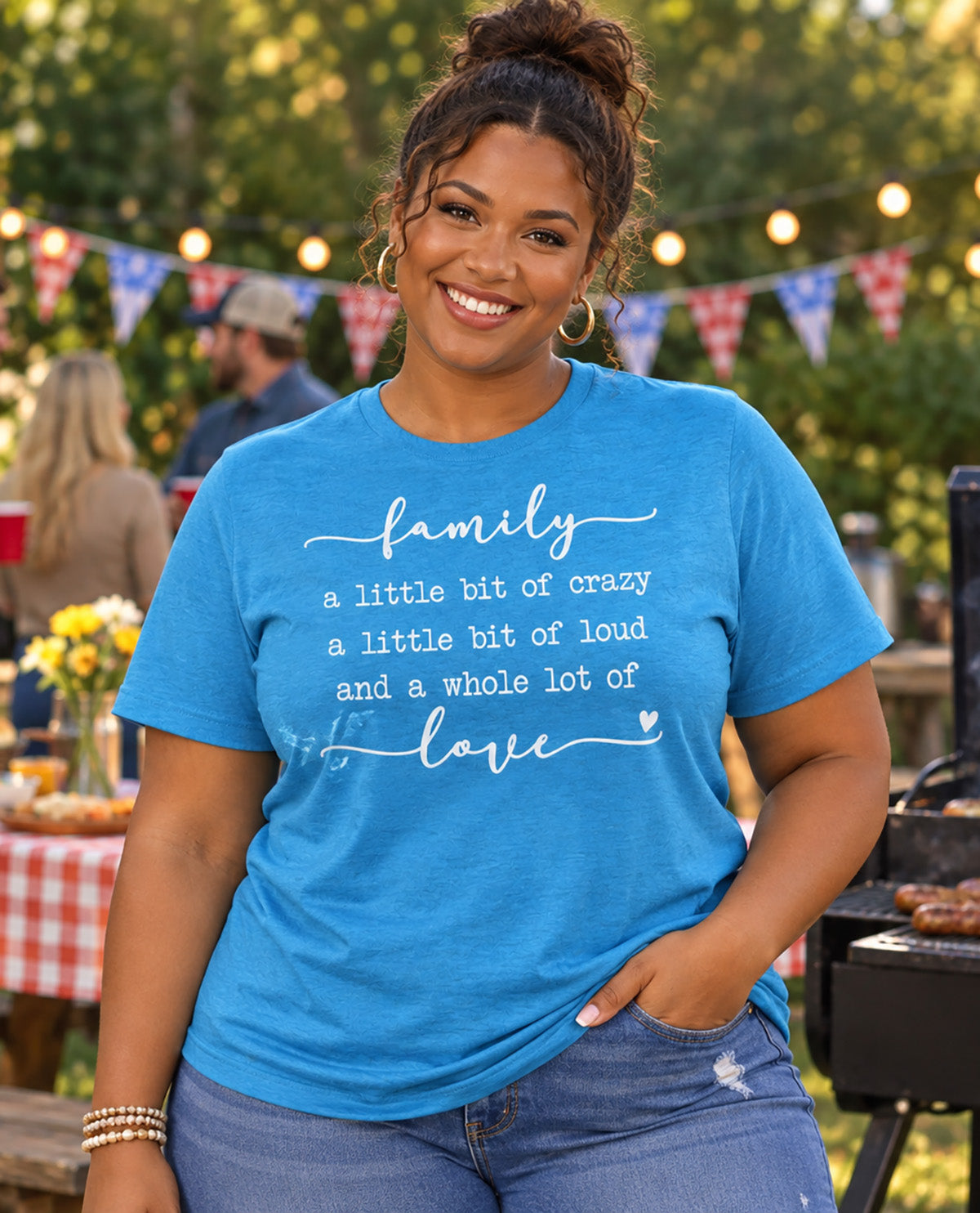 A woman wearing a bright blue graphic t-shirt with the text 'family a little bit of crazy a little bit of loud and a whole lot of love' in white script and print, set against an outdoor backyard party background.