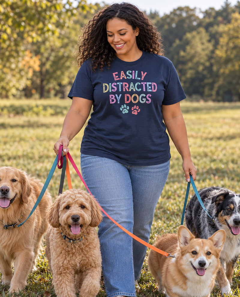 A woman walking outdoors in a grassy park with four dogs, wearing a navy blue graphic t-shirt that reads 'EASILY DISTRACTED BY DOGS' in colorful lettering with paw prints.