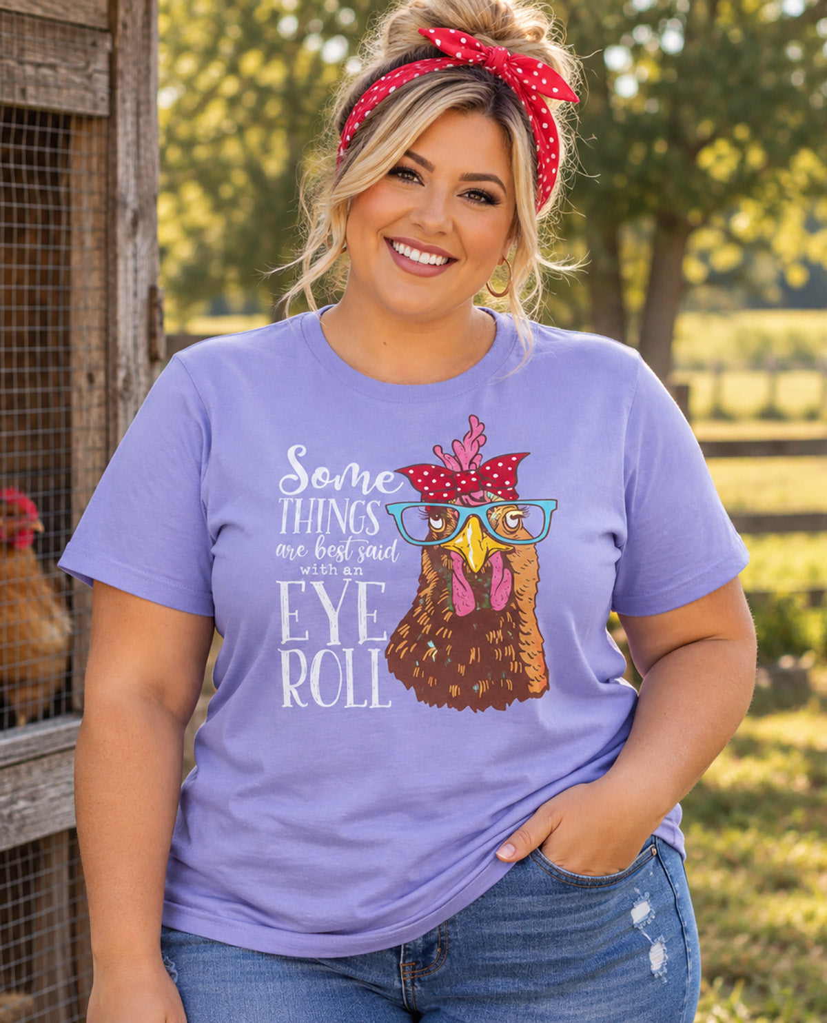 A woman wearing a light purple graphic t-shirt with a cartoon chicken wearing glasses and a red polka-dot headband, standing outdoors in front of a rustic wooden coop.