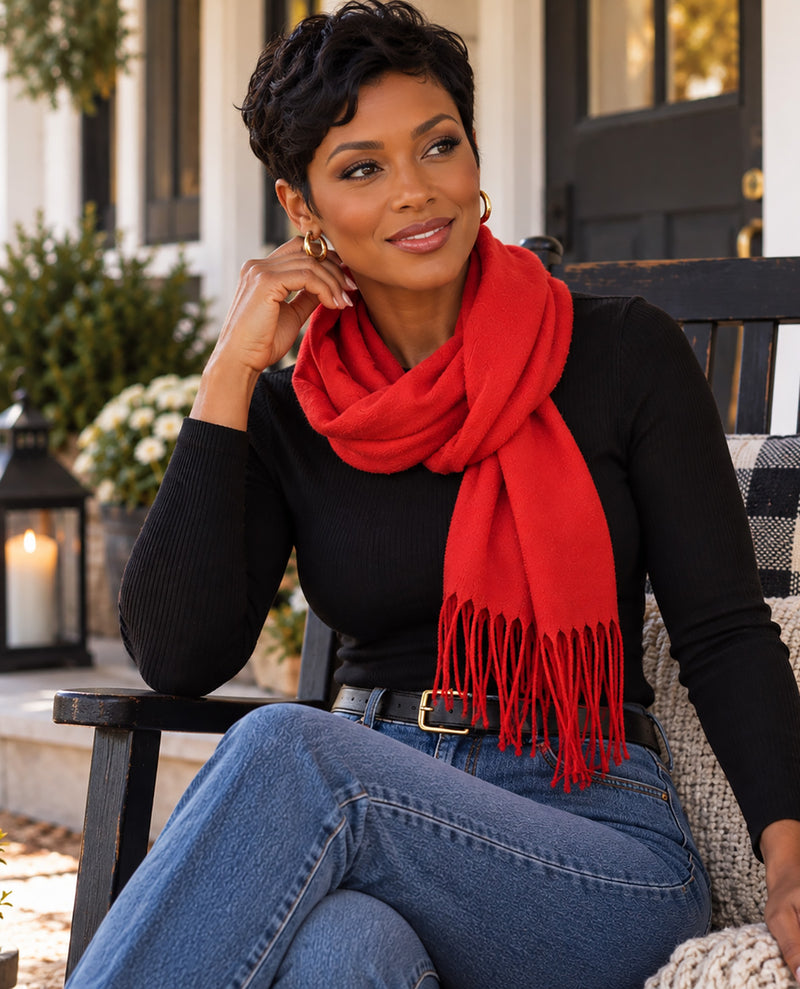 A woman sitting on a porch chair wearing a bright red knit scarf with fringe, a black long-sleeve top, and blue jeans.