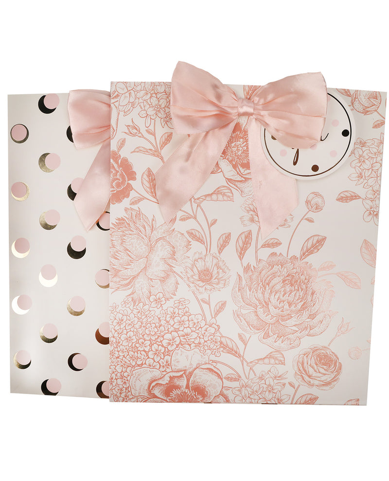 A pink and white floral gift bag with a large satin bow on top, a round white gift tag, and a matching white pouch with gold and pink moon cutouts behind it, shown on a white background.