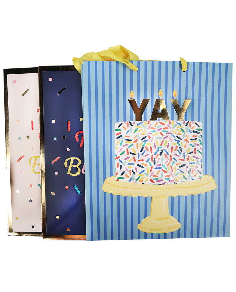 Three birthday gift bags with party-themed designs, including a blue striped bag with a birthday cake and candles, a navy bag with colorful confetti and letters, and a light pink bag with confetti and partial lettering.