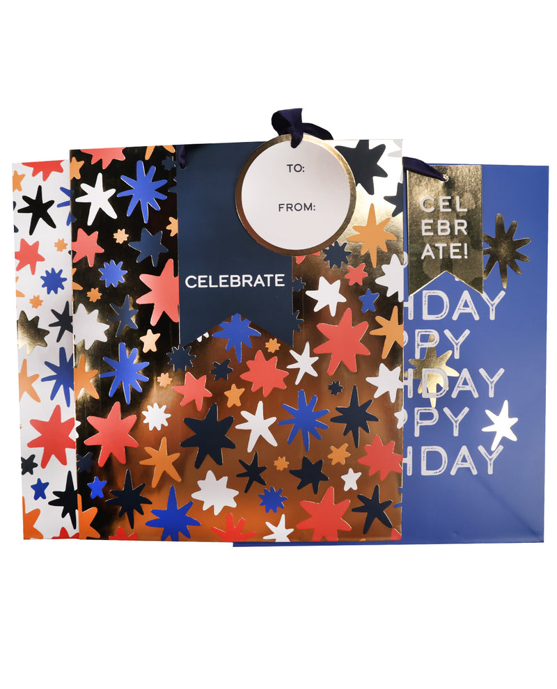 A set of patriotic star-patterned gift bags and a matching gift tag on a white background. The bags feature colorful metallic star cutouts in red, orange, blue, black, and white over dark navy, gold, and blue backgrounds.