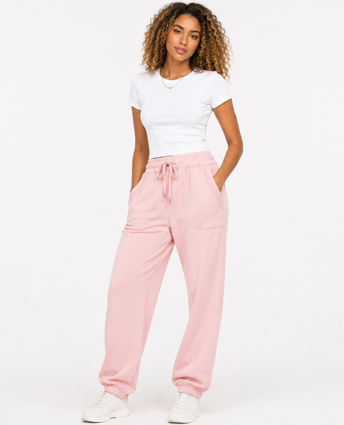 A woman modeling a pair of light pink drawstring sweatpants with a white t-shirt and white sneakers, standing against a plain white studio background.
