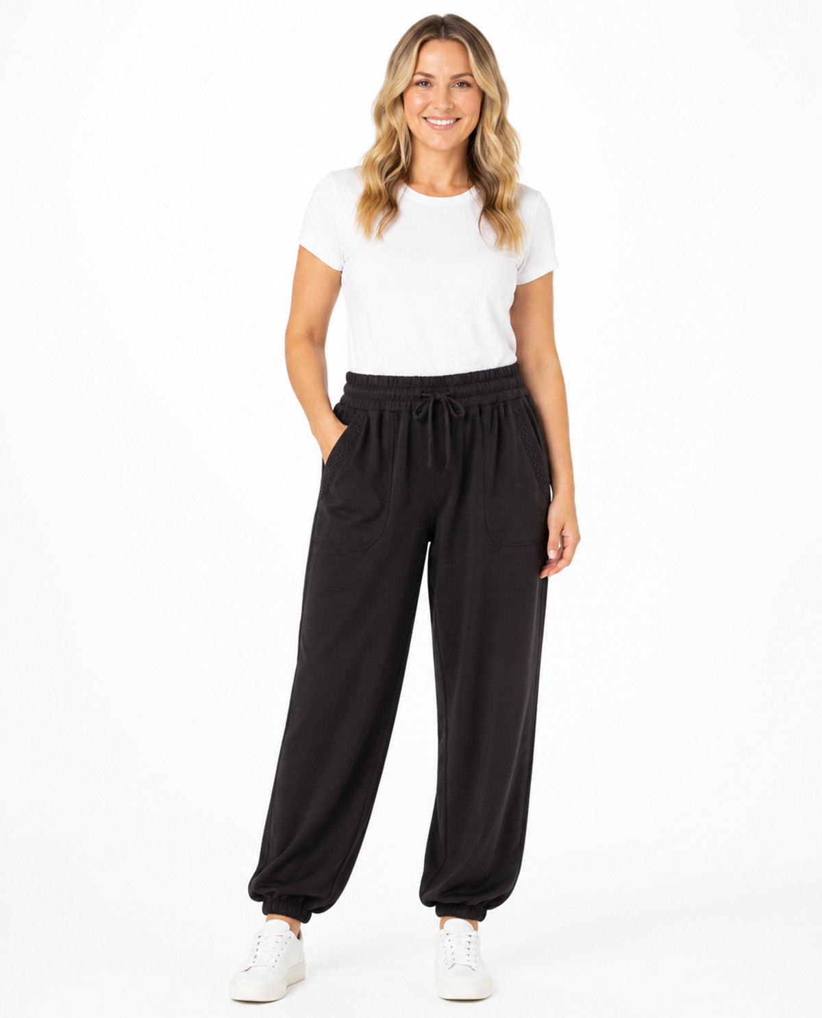 A woman modeling black wide-leg drawstring sweatpants with a white t-shirt and white sneakers, standing against a solid white studio background.