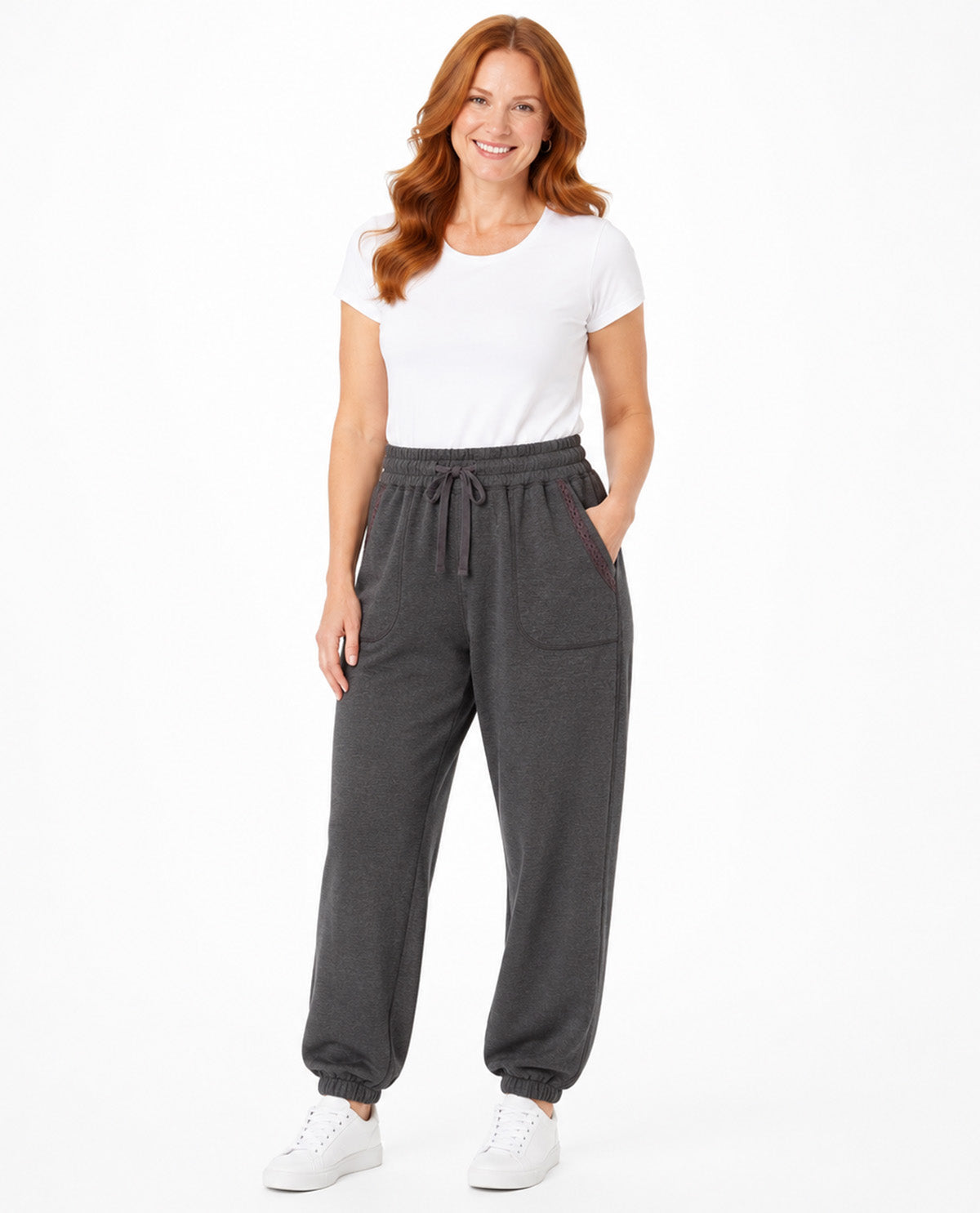 A woman modeling dark gray jogger sweatpants with a drawstring waist and side pockets, paired with a white t-shirt and white sneakers, against a plain white background.