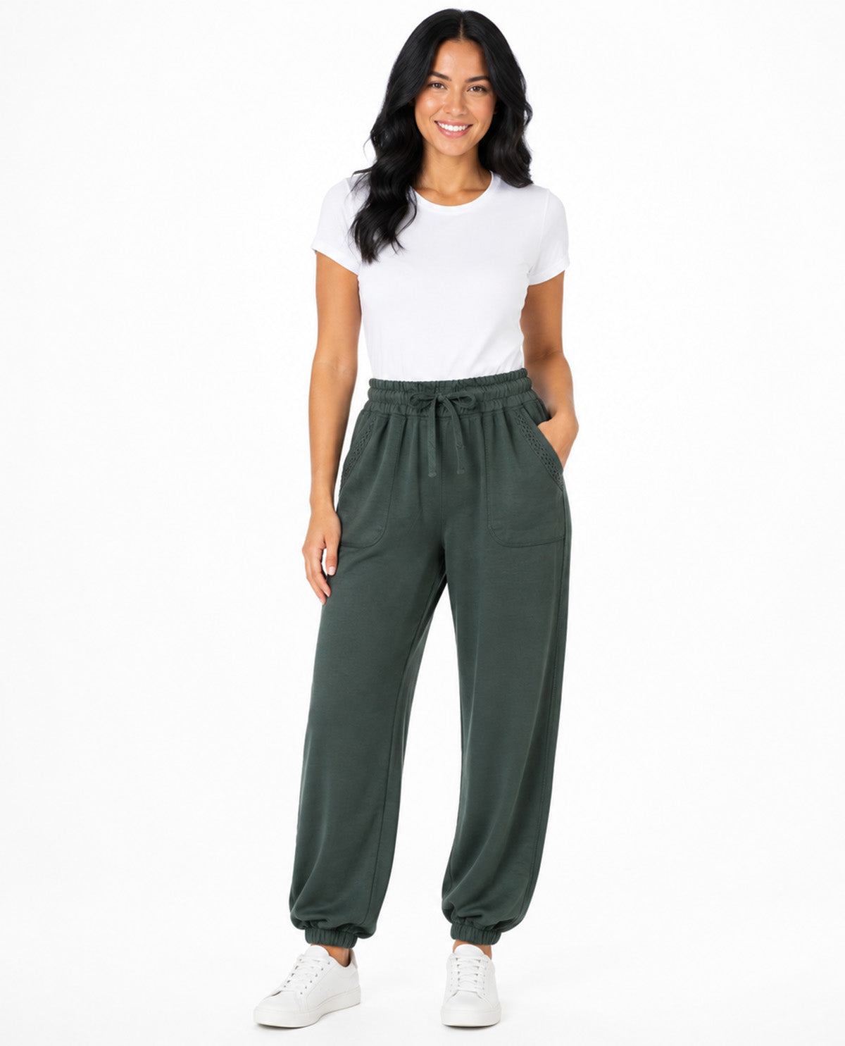 A woman modeling dark green drawstring sweatpants with a white t-shirt and white sneakers, standing against a solid white background.