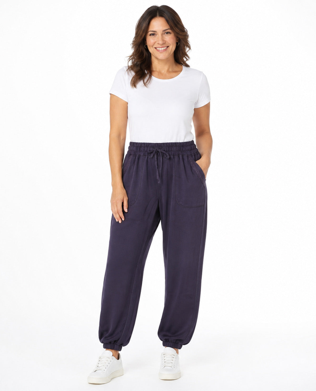 A woman modeling a pair of dark purple drawstring jogger pants with a white t-shirt and white sneakers, standing against a plain white studio background.