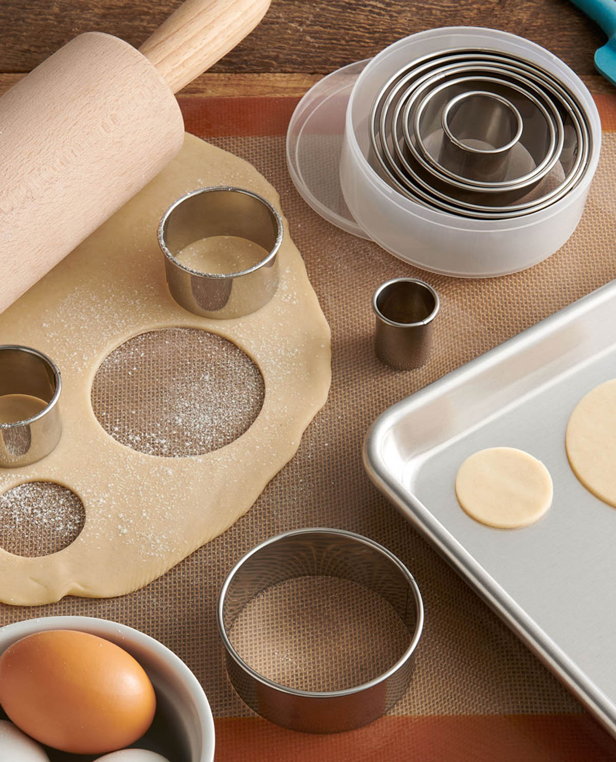 A set of round metal and plastic cookie cutters arranged on dough and a baking tray, with a wooden rolling pin, eggs, and a baking mat in a kitchen-style scene.