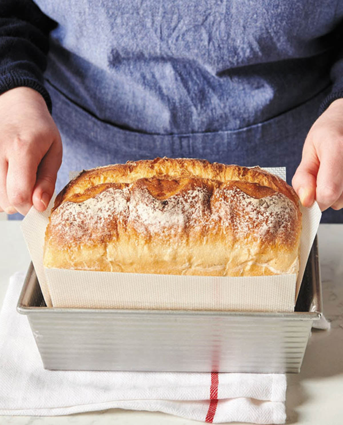 Loaf of bread in a baking pan held by a person wearing a blue apron.