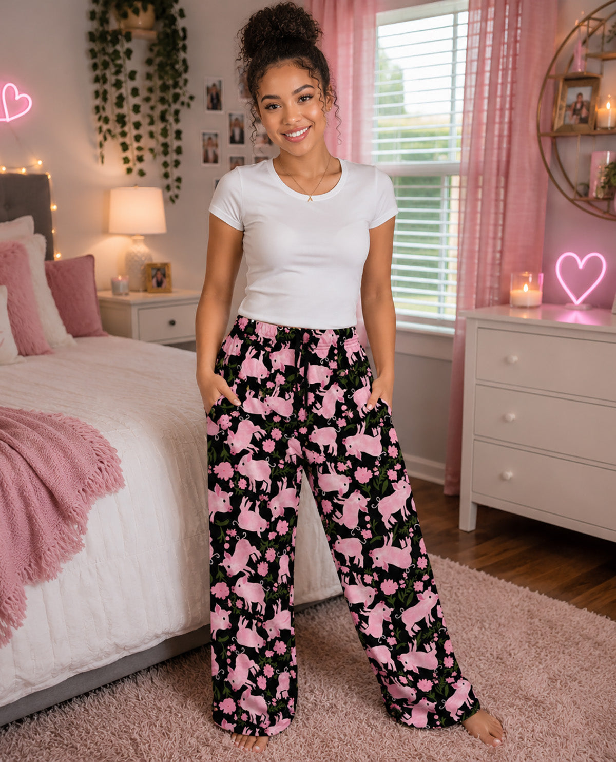 A woman stands barefoot in a bedroom wearing black wide-leg pajama pants with a pink pig print and a white t-shirt. The room features pink accents, including curtains, a rug, and a bed.