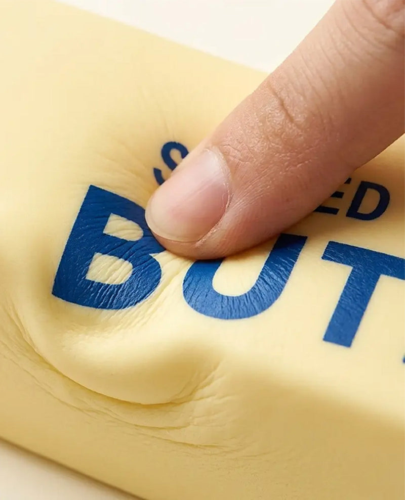 A close-up of a finger pressing into a pale yellow, squishy material printed with bold blue text reading 'BUT'.