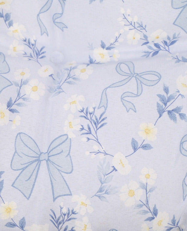 A close-up of a light lavender fabric with a repeating pattern of pale blue bows, blue leaves, and small white flowers with yellow centers.