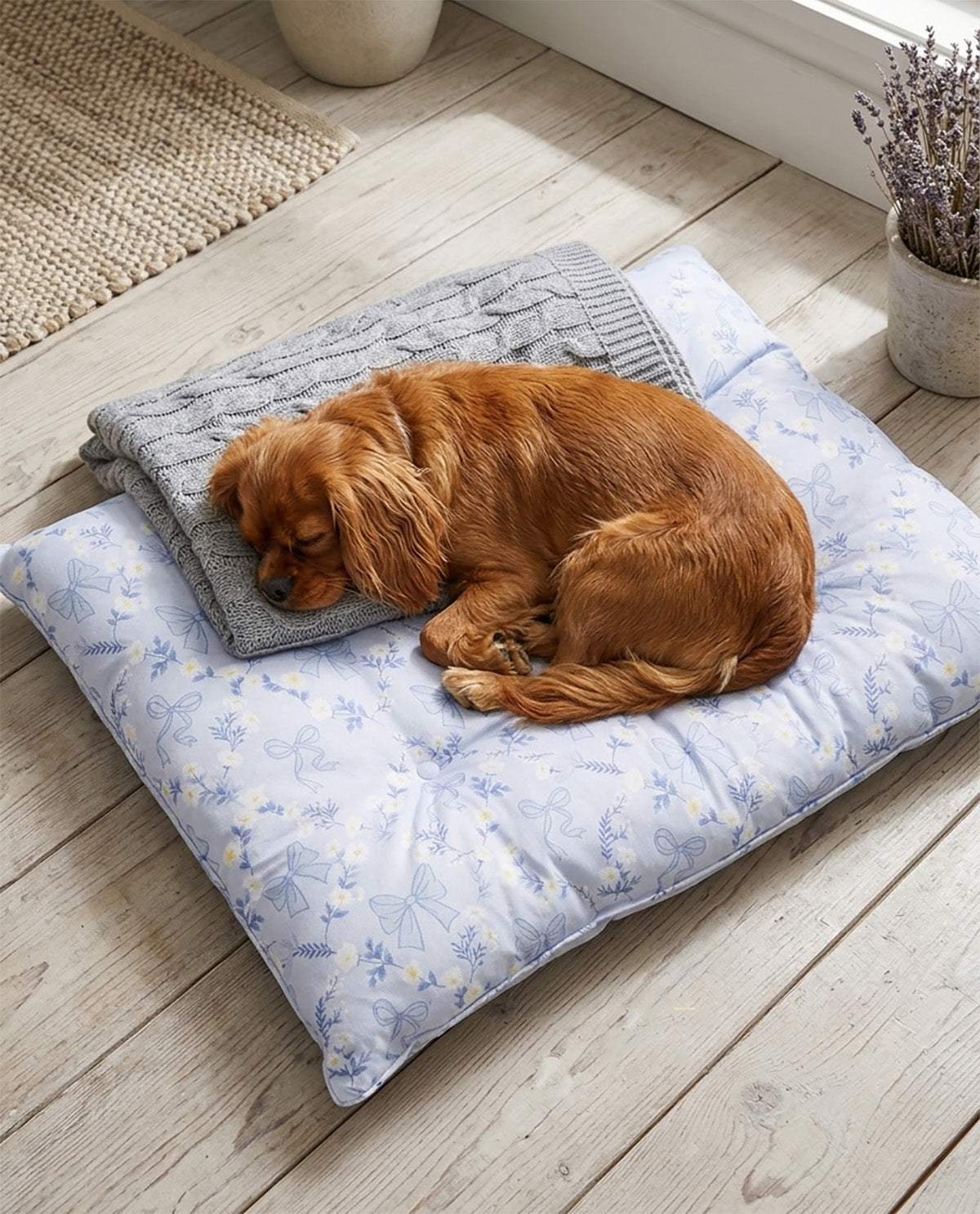 A golden retriever dog sleeps curled up on a square light blue floral pet bed with a folded gray knit blanket underneath, placed on a light wood floor in a bright room.