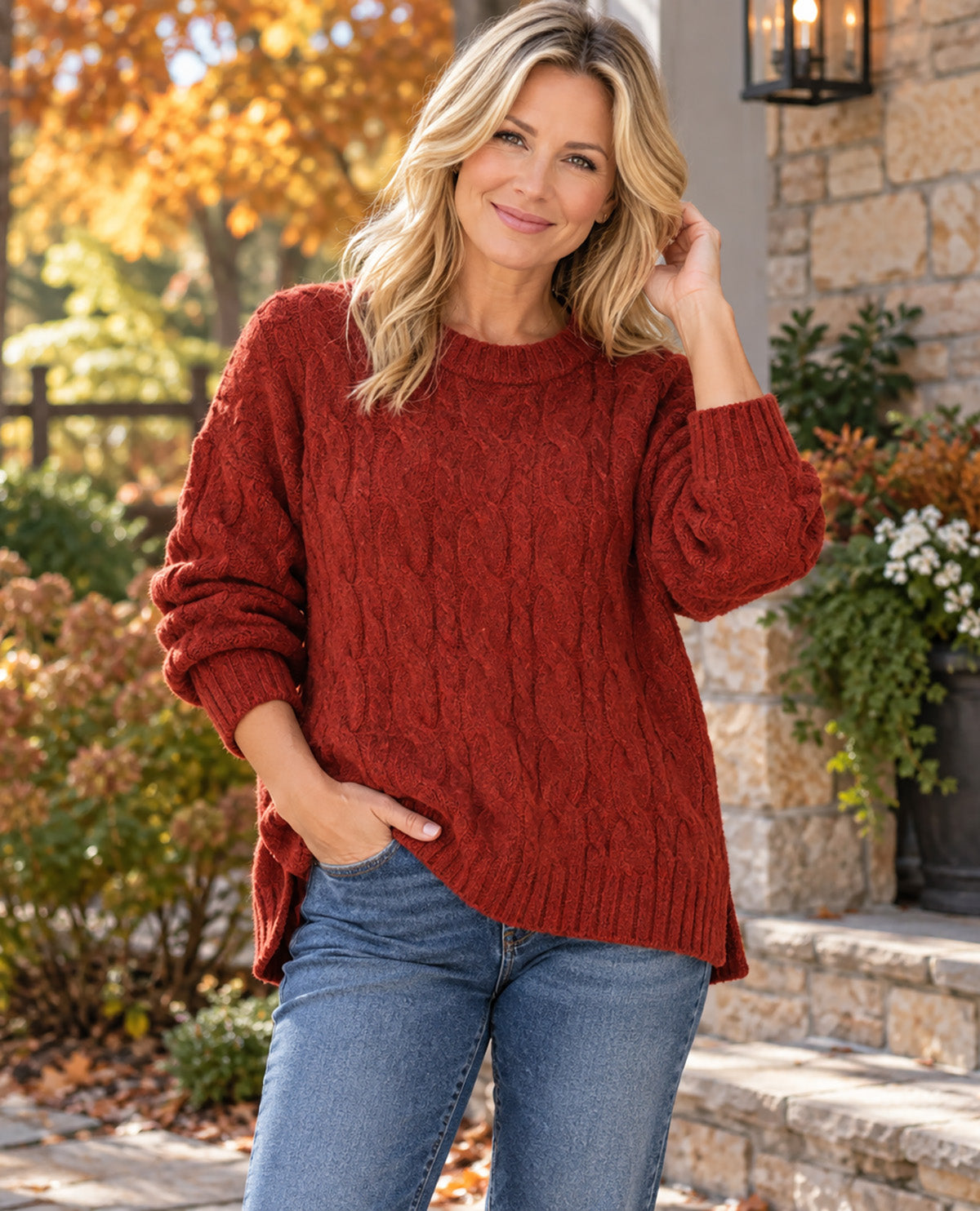 A woman wearing a rust-red cable-knit sweater and blue jeans, standing outdoors in front of a stone wall with autumn foliage.