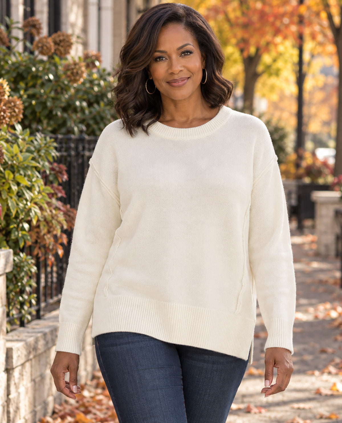 A woman wearing a cream-colored knit crewneck sweater and dark blue jeans, standing on an outdoor city sidewalk with autumn trees in the background.