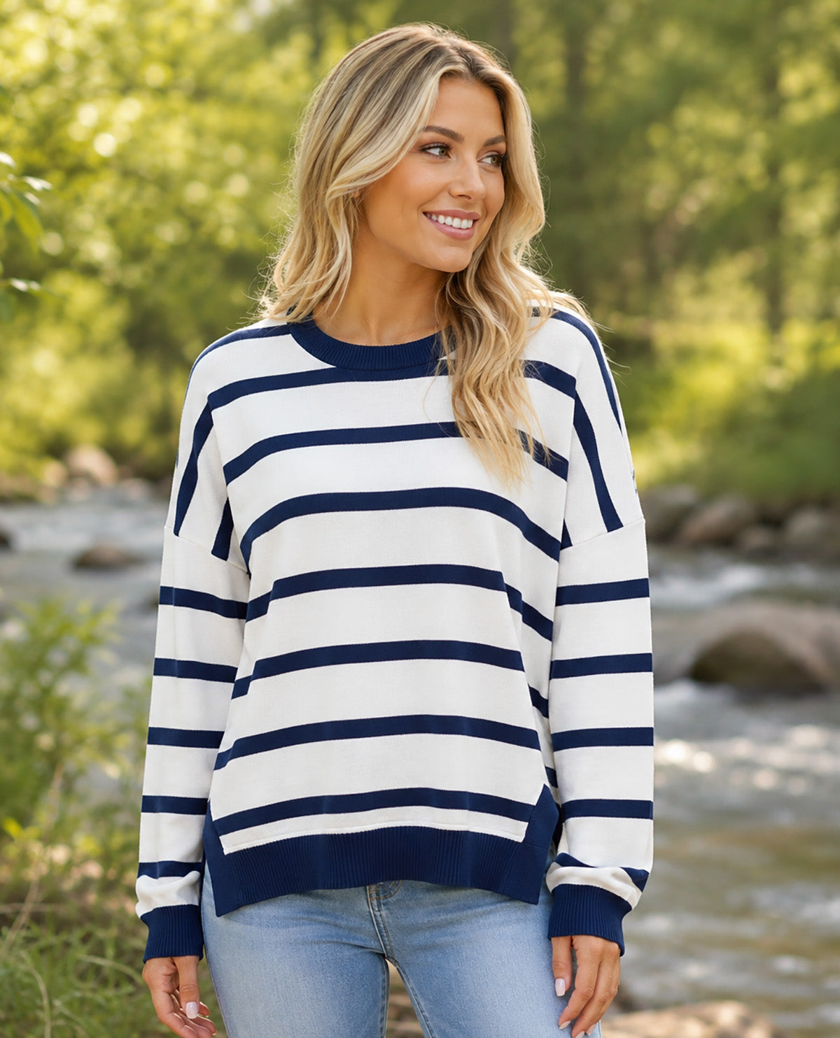 A woman wearing a navy blue and white horizontally striped long-sleeve sweater, paired with light blue jeans, standing outdoors in front of a blurred background of green trees and a flowing river.