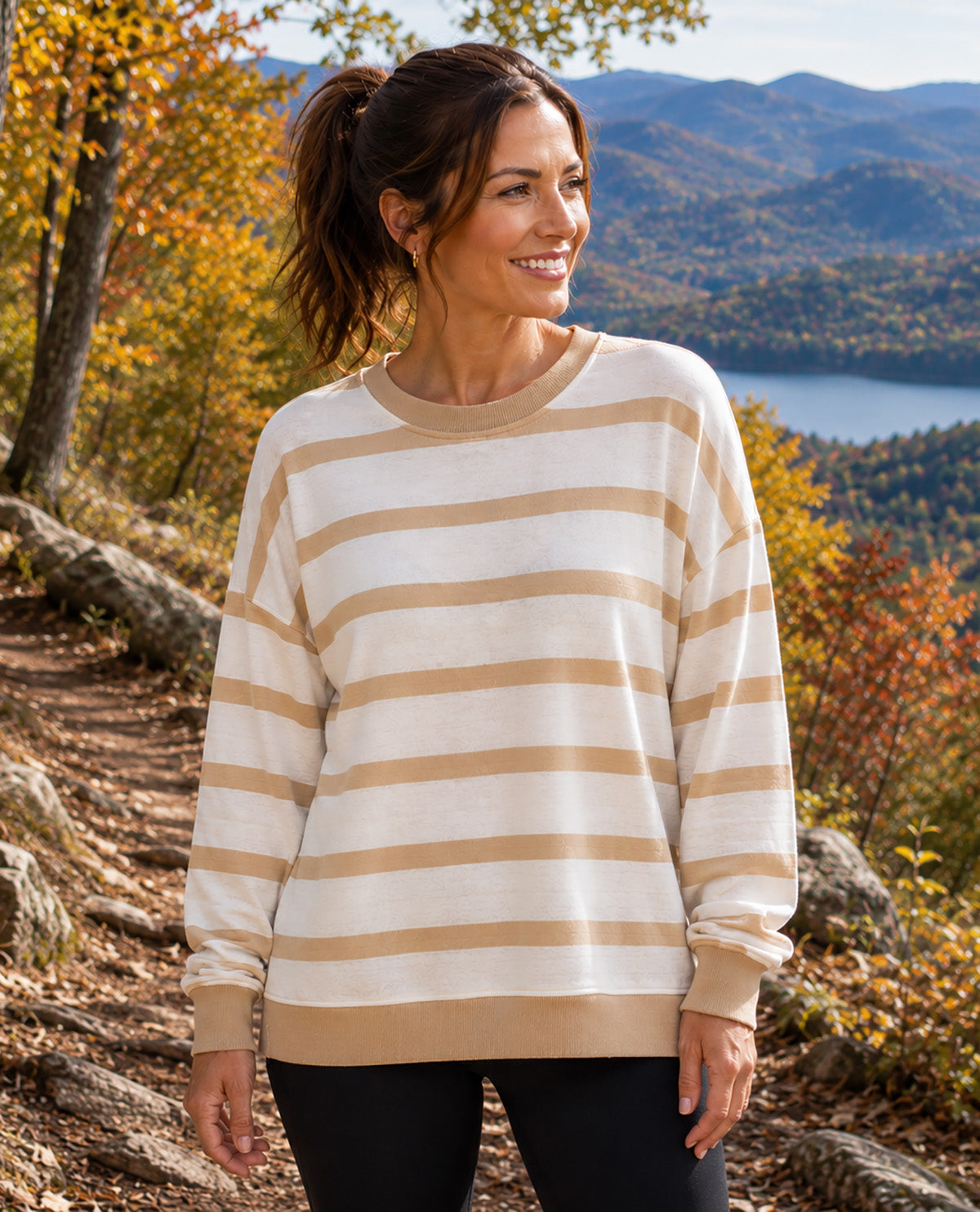 A woman wearing a cream and tan horizontally striped crewneck sweatshirt, standing on an outdoor mountain trail with autumn foliage and a lake in the background.