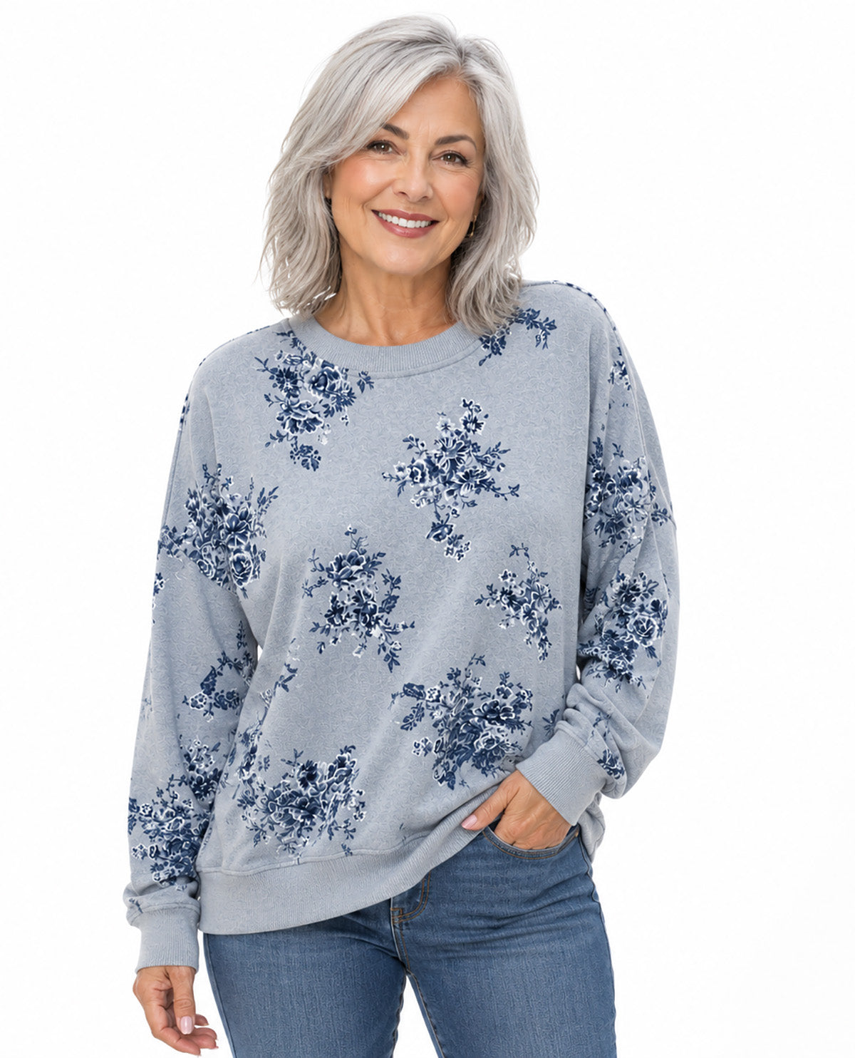 A smiling woman with shoulder-length gray hair wears a light gray crewneck sweatshirt with a navy blue floral print, paired with blue jeans, against a plain white background.