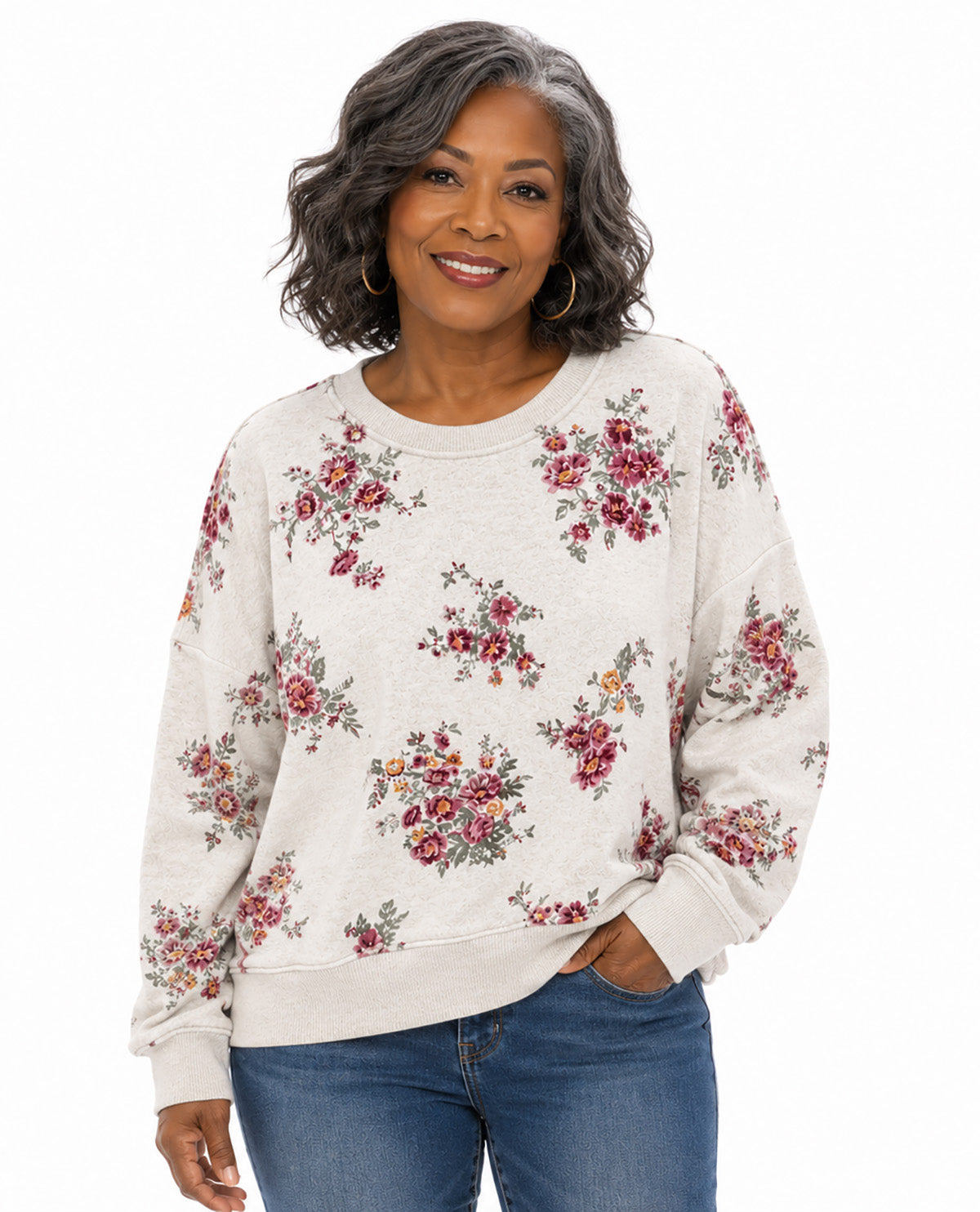 A woman wearing a cream-colored crewneck sweatshirt with a red and green floral pattern, paired with blue jeans, against a plain white background.