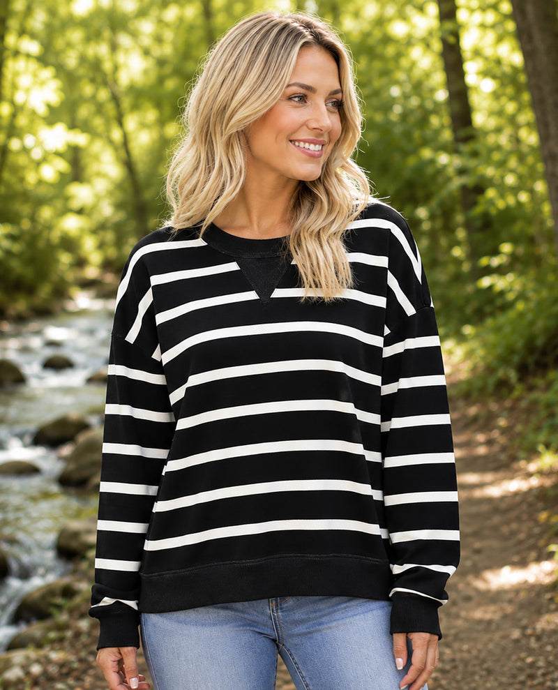 A woman wearing a black and white horizontally striped long-sleeve crewneck sweatshirt, paired with light blue jeans, standing outdoors in a sunlit wooded area near a stream.