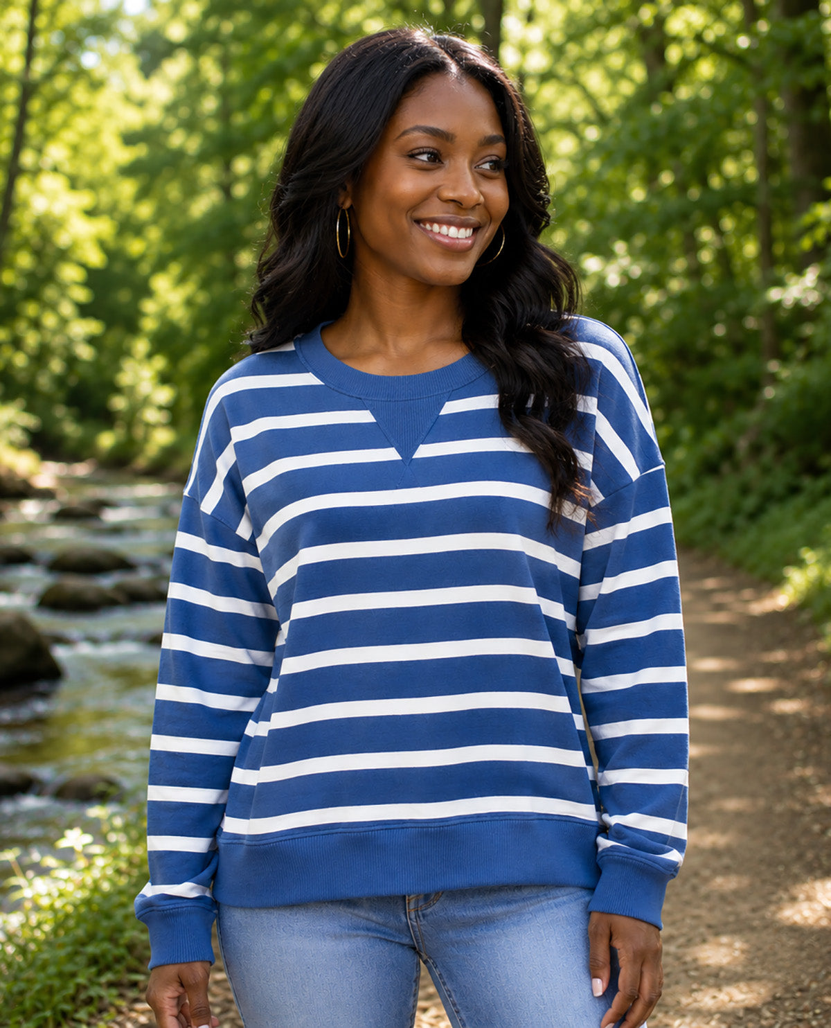 A woman wearing a blue and white horizontally striped crewneck sweatshirt, standing outdoors on a dirt path with a stream and green trees in the background.