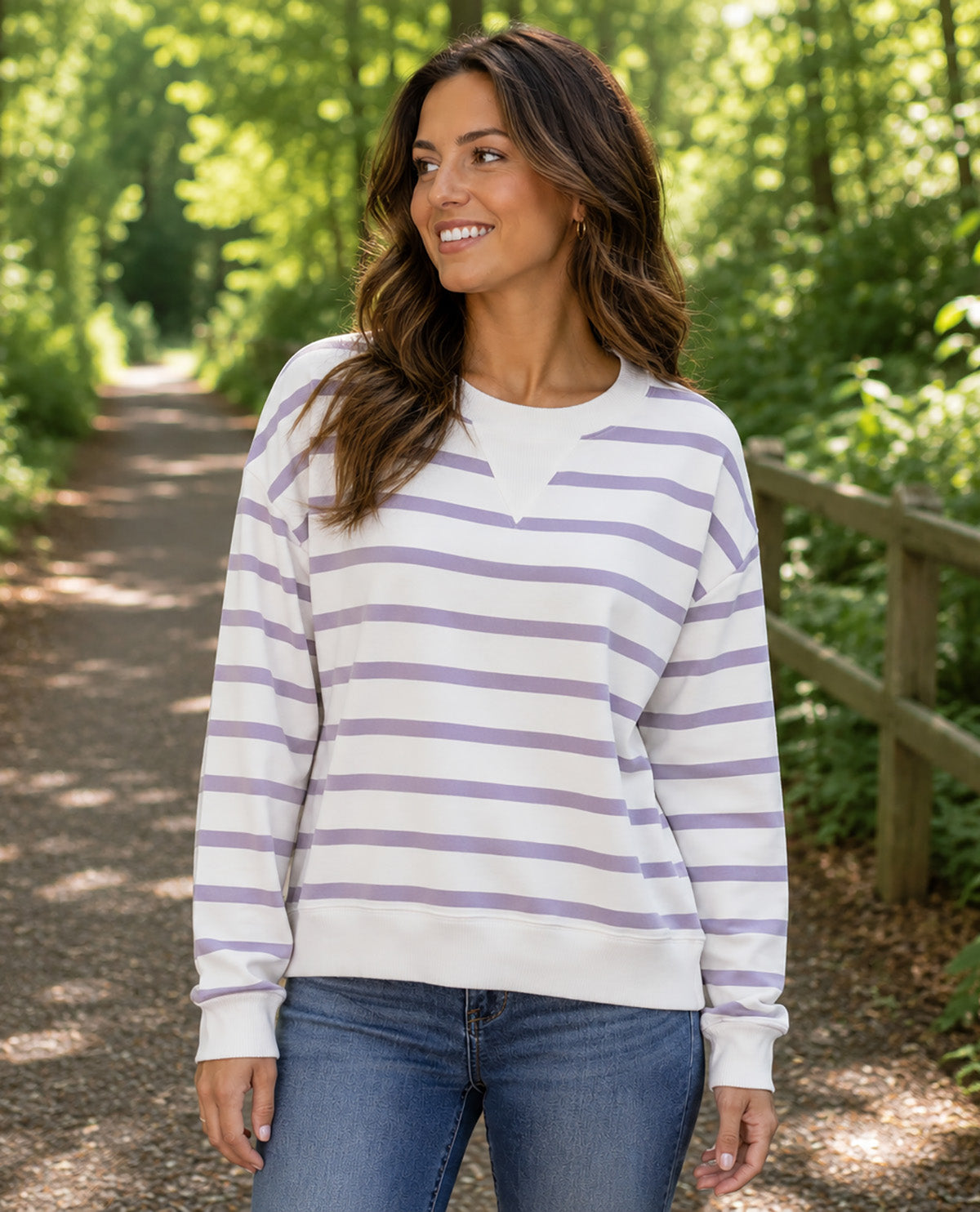 A woman wearing a white long-sleeve crewneck sweatshirt with horizontal lavender-purple stripes, paired with blue jeans, standing on a sun-dappled forest path.