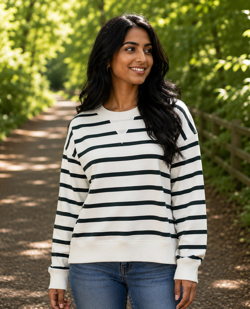 A woman wearing a black and white horizontally striped long-sleeve crewneck sweatshirt, paired with blue jeans, standing on a sun-dappled park path.
