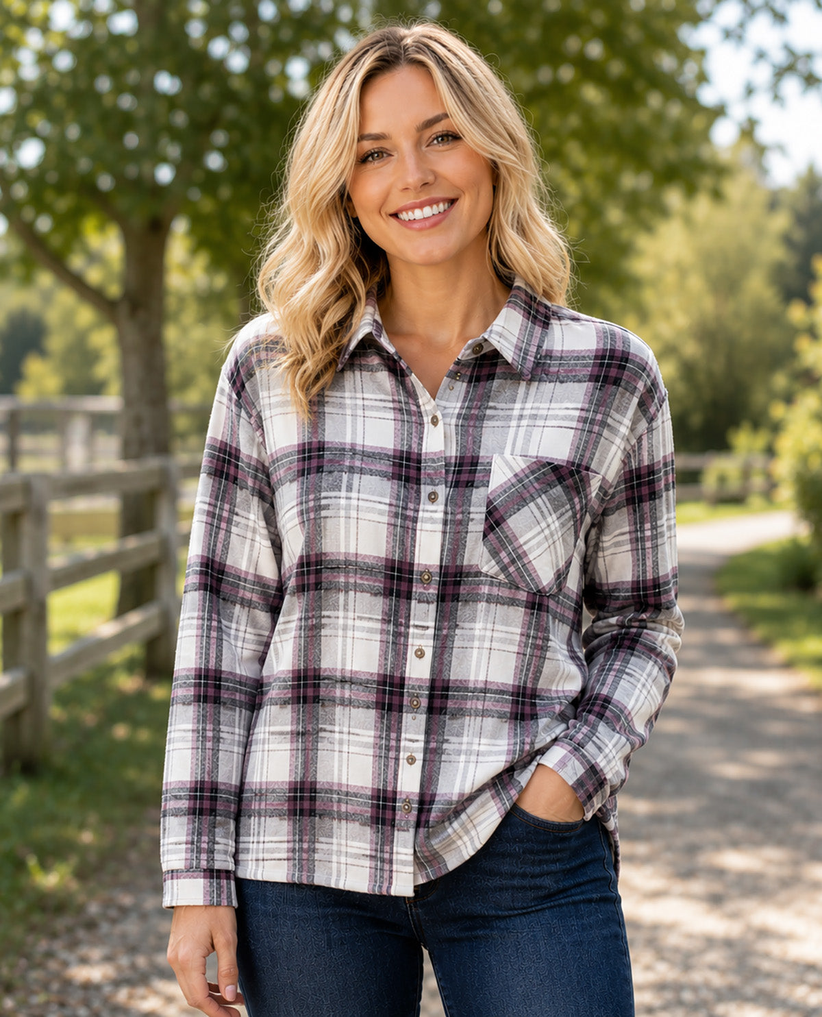 A woman wearing a long-sleeve plaid button-up shirt in white, gray, and dark purple, paired with dark blue jeans, standing outdoors on a dirt path with trees and a wooden fence in the background.