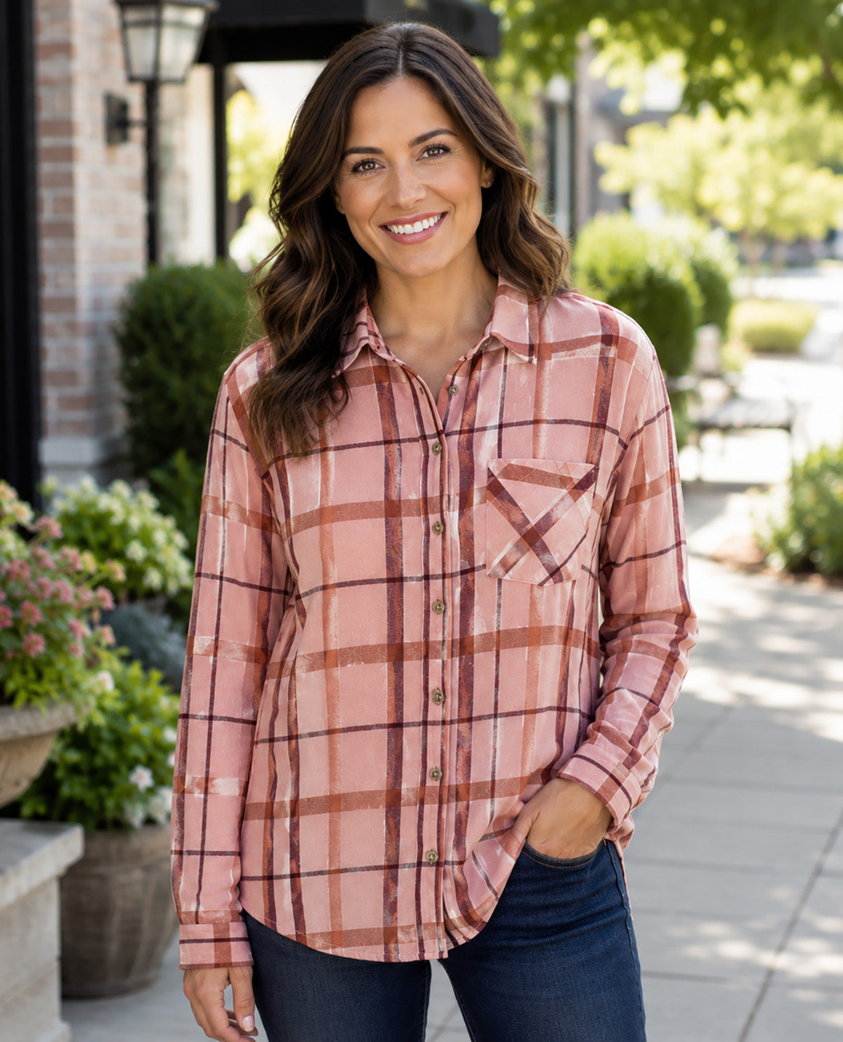 A woman smiling outdoors while wearing a long-sleeve button-up plaid shirt in dusty pink and brown tones, paired with dark blue jeans.