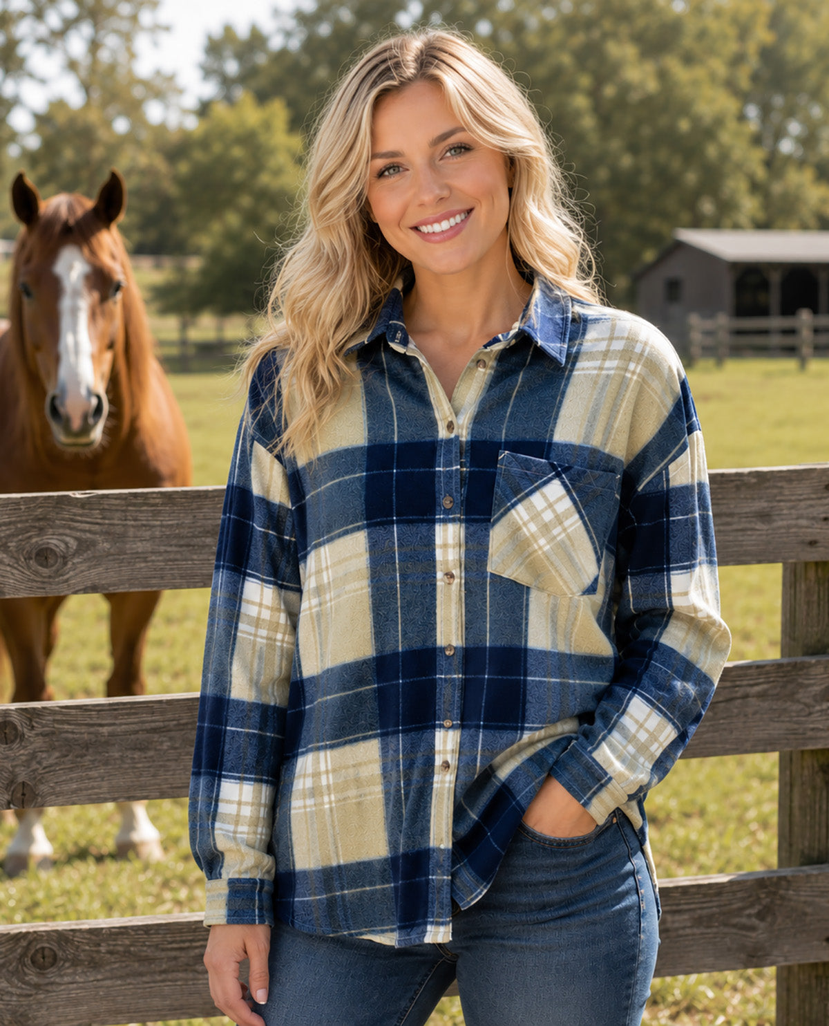 A woman stands outdoors in front of a wooden fence wearing a long-sleeve plaid button-up shirt in navy blue and cream, paired with blue jeans. A brown horse is visible in the background on the left, with trees and a small building behind her.