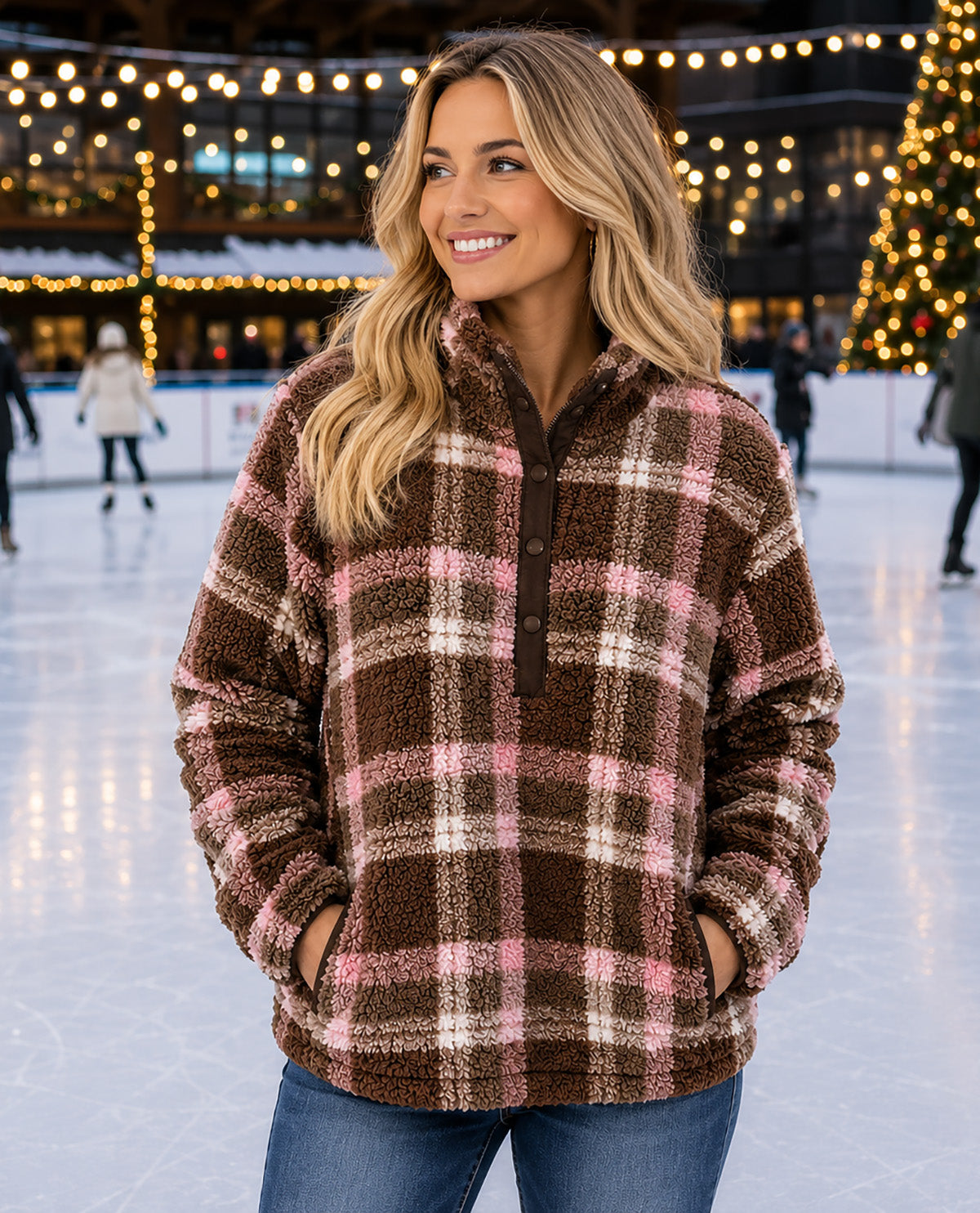 A woman wearing a brown, pink, and white plaid fleece pullover jacket stands on an ice rink at night with holiday string lights in the background.