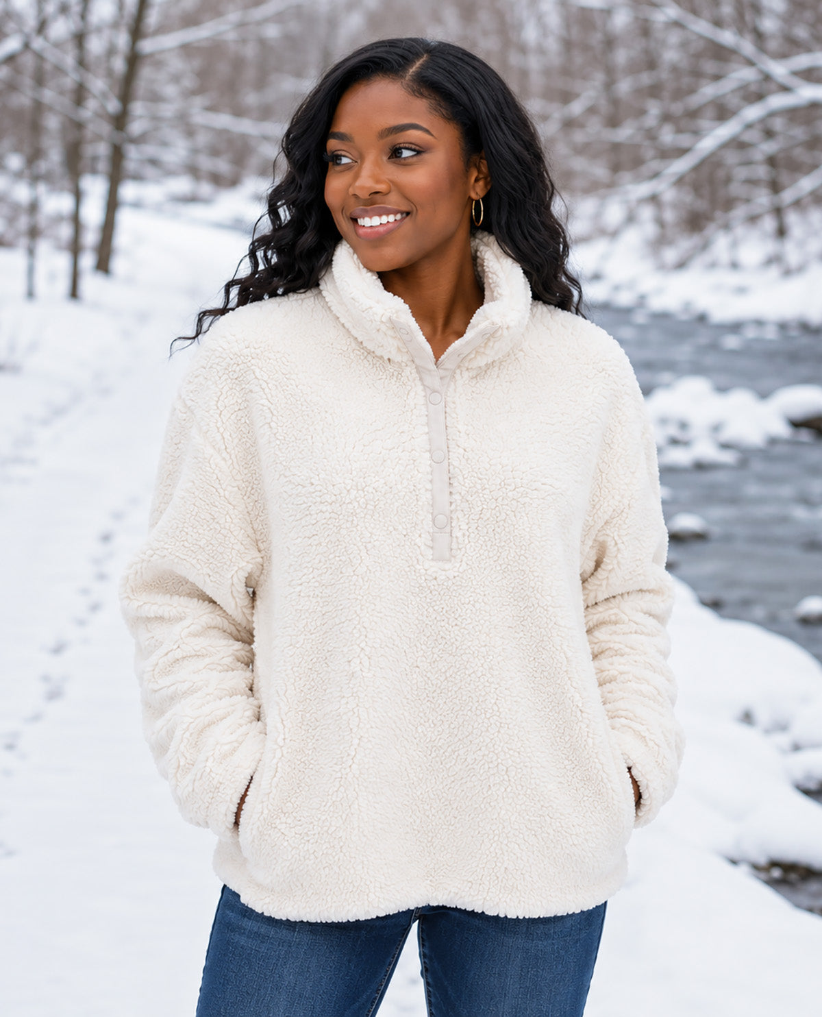 A woman wearing a cream sherpa fleece half-zip pullover sweater with a high collar, standing in a snowy outdoor setting next to a stream.