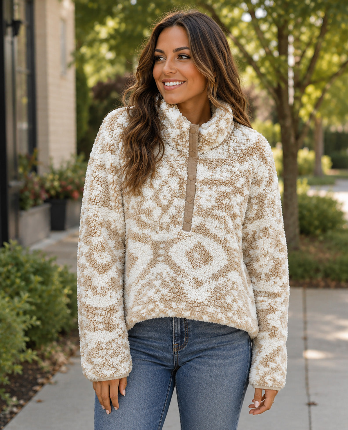 A woman wearing a cream and tan patterned quarter-zip fleece pullover sweater with blue jeans, standing outdoors on a sidewalk with trees and a building in the background.