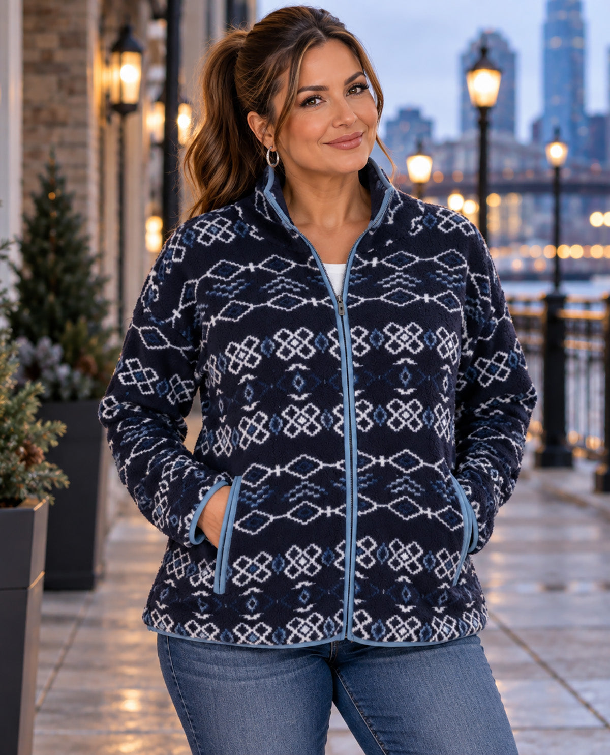 A woman wearing a navy blue fleece jacket with a white geometric pattern and light blue trim, standing on an outdoor walkway at dusk.