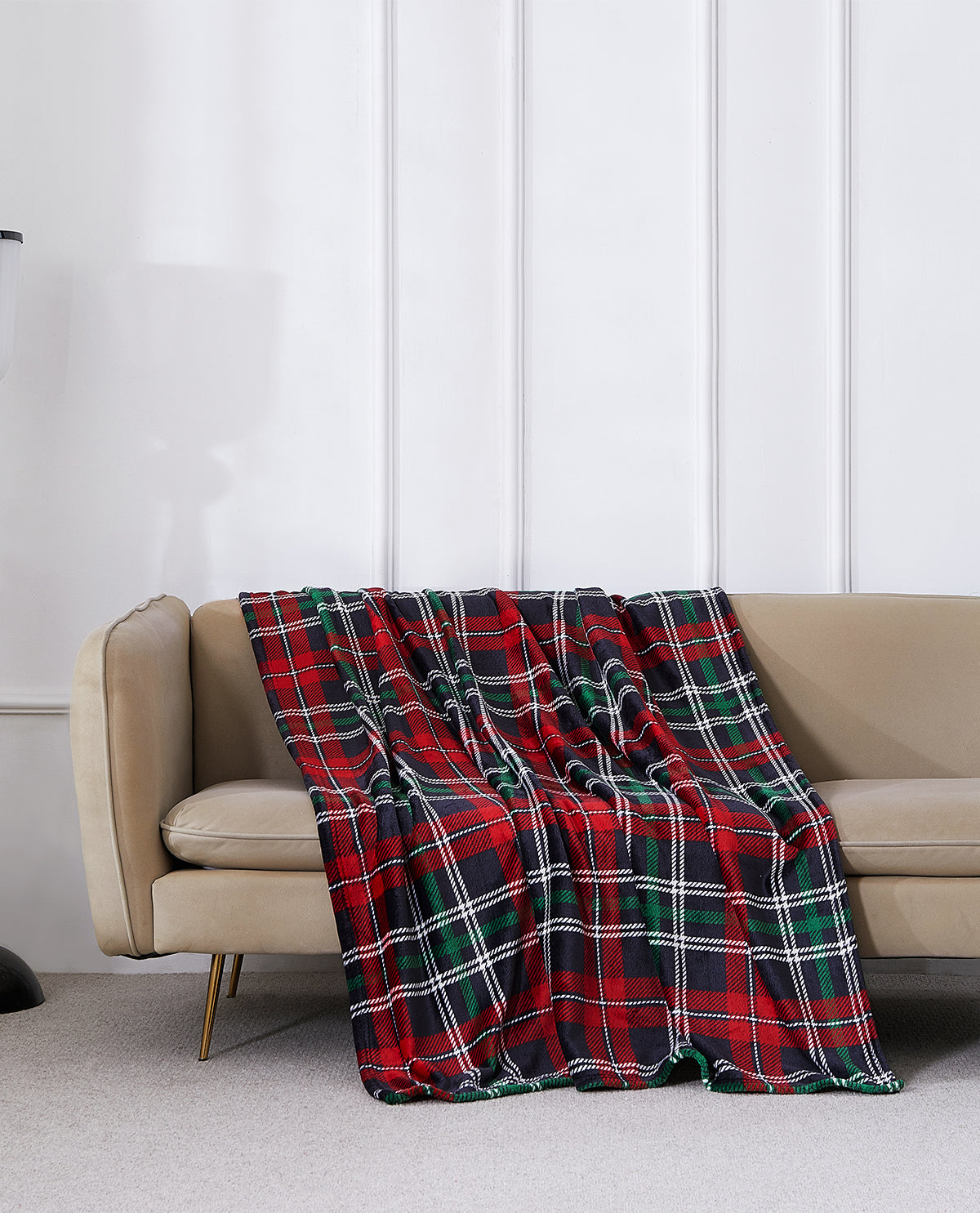 A red, green, and black plaid throw blanket is draped over a beige sofa in a modern living room setting with a white paneled wall.