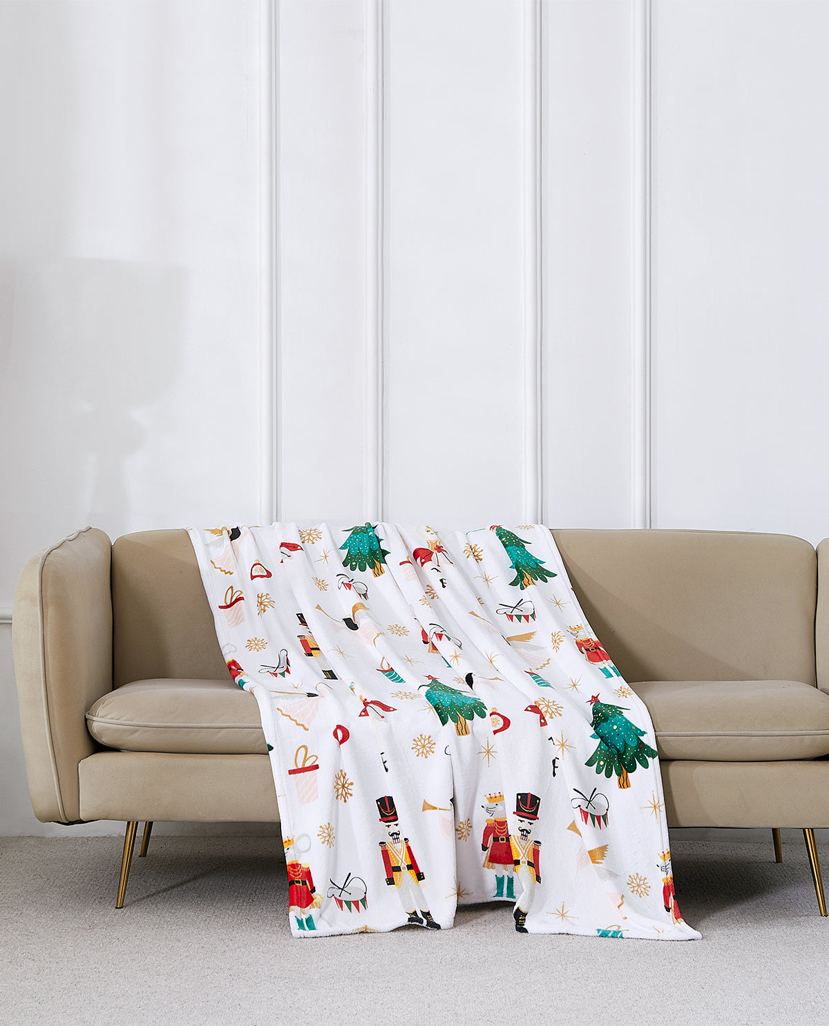 A white throw blanket with a colorful Christmas nutcracker and tree pattern is draped over a beige sofa in a modern living room setting.
